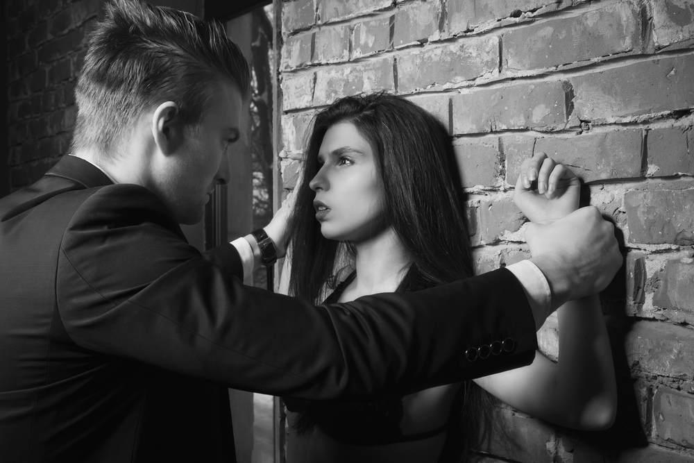 Man pinning a woman's wrists against a brick wall.