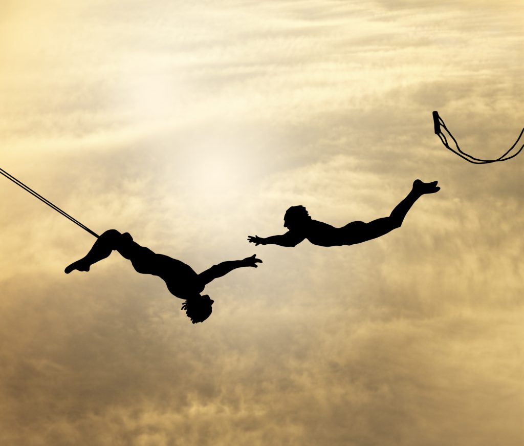 Two trapeze artists. One has their legs hooked around the swing while the other has leapt off of their swing and is in midair about to grab the hands of the other person.